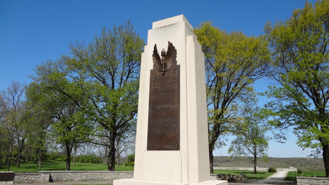 A tall gray stone monolith with a brown bronze plaque affixed to the front with trees in background