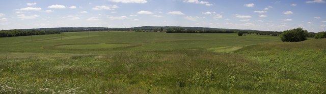 A green, grassy field with taller areas of uncut grass in geometric shapes under a partly cloudy sky