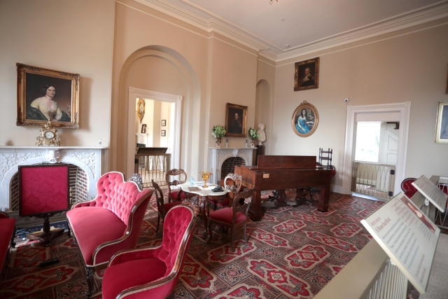 Portraits hang on the walls of the parlor room with red velvet furniture and a small dining table.