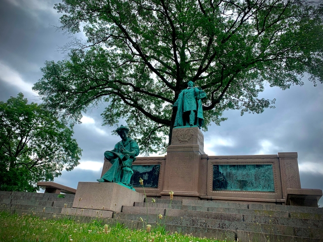 A statue of a young and an adult Samuel Colt in front of a tree with a cloudy sky.
