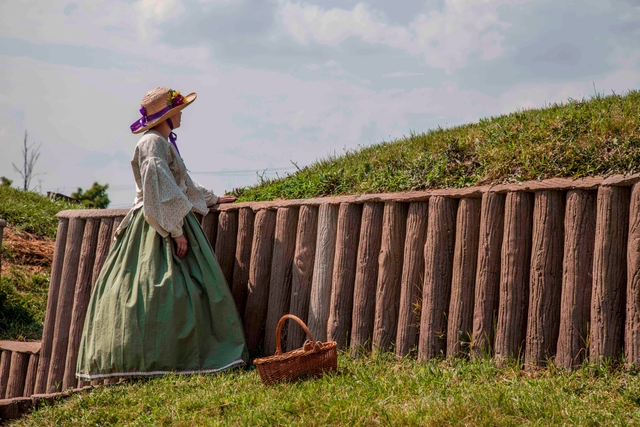 A woman in a 19th-century dress stands along an earthwork fortification