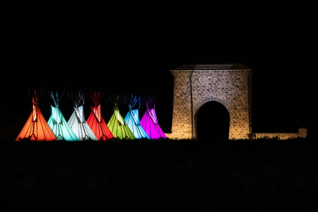Multi-colored teepees are lit in front of a large, stone arch under the black night sky.