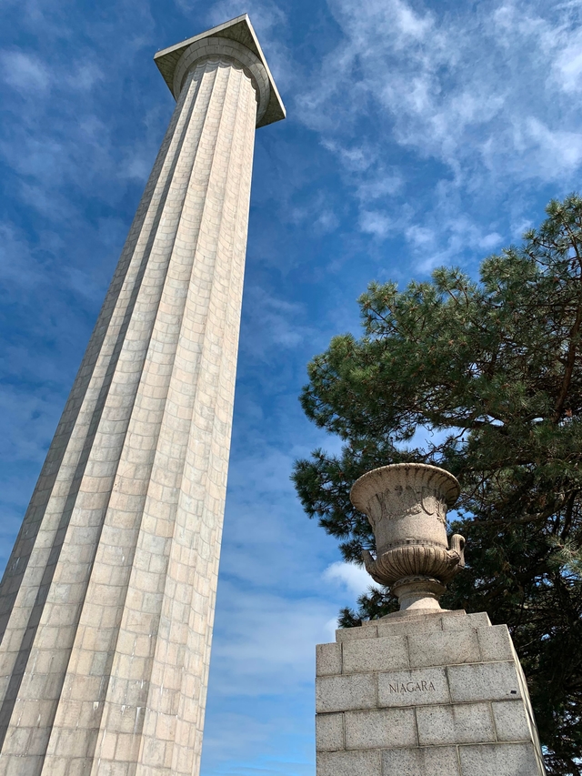 352 foot tall stone memorial column rises up to touch blue sky. In front a 5' urn on pedestal