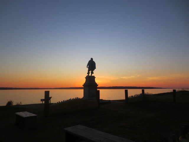 Statue on a pedestal silhouetted against a sunset over a river