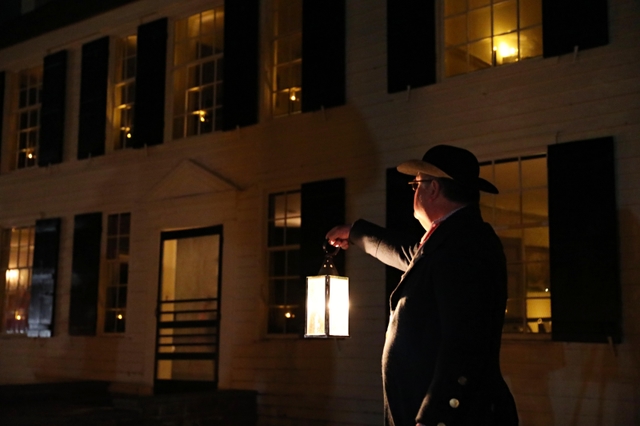 A man in 18th-century style clothing holds a candlelit lantern in front of a 2-story yellow house.