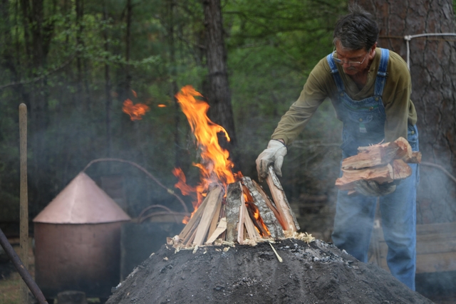 Man loading wood on to a dirt mound with a fire burning.