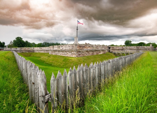 Puffy white clouds billow over log cabin-like walls. An American flag flies on a wooden pole