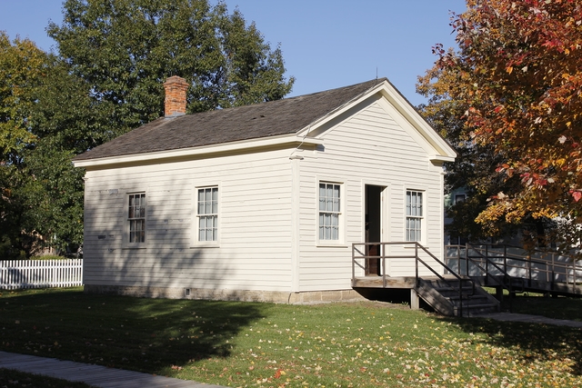 A one-story wooden building painted off-white has two windows and a central doorway.