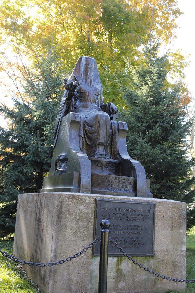 A cast bronze figure of a veiled goddess sits on a throne mounted on a concrete pedestal.