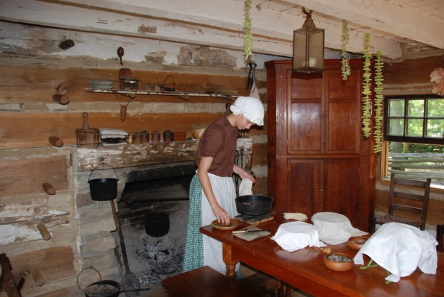 Park ranger is period dress standing near fireplace, cooking in the cabin.