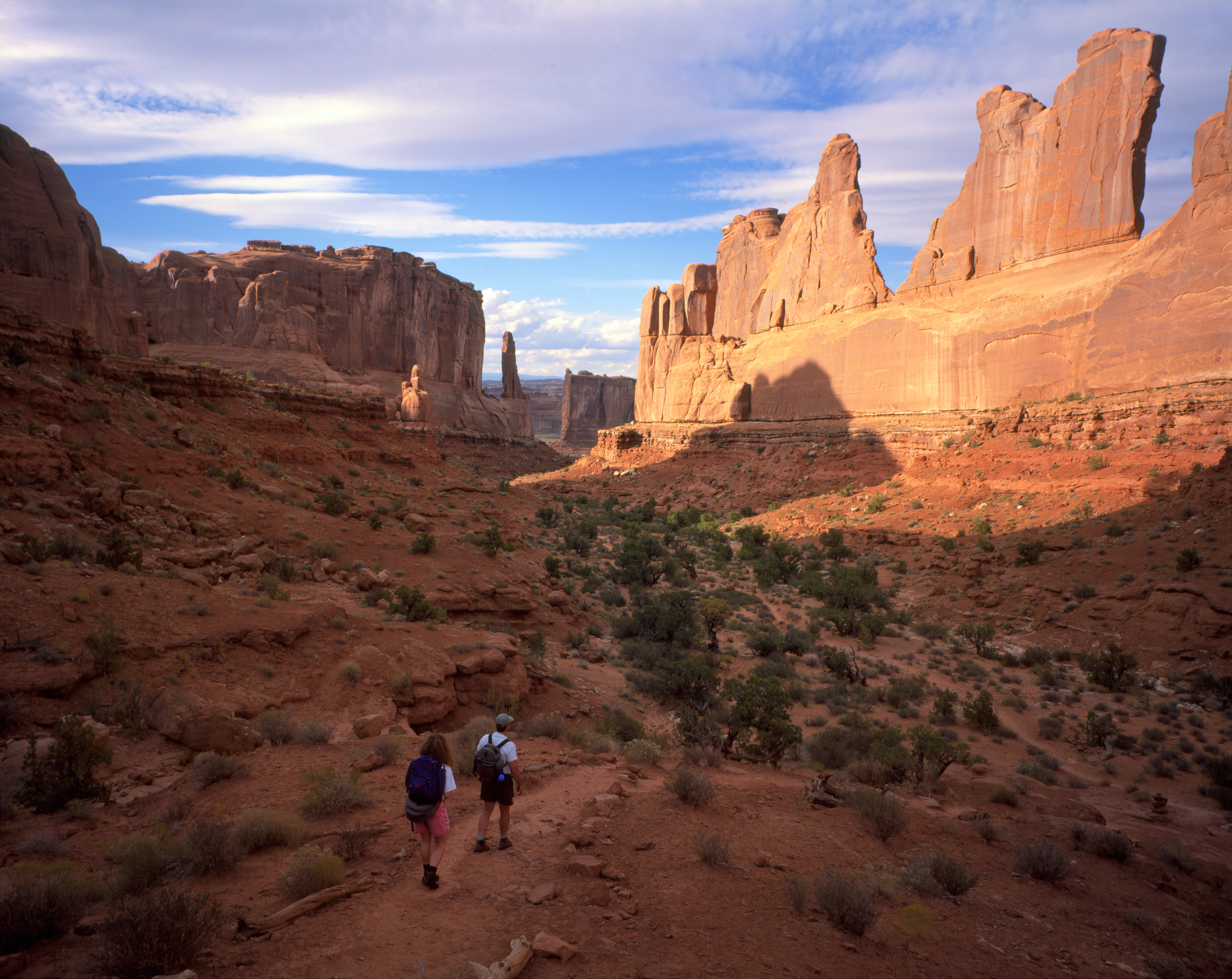 two hikers descend a broad wash with tall rock walls on either side.