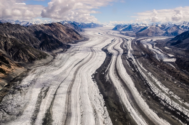 A large glacier with stripes of different colored rock nestled in between barren mountain slopes.