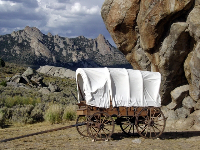 A white canvas wagon sits in front of a large rock buttress with mountains in the distance.