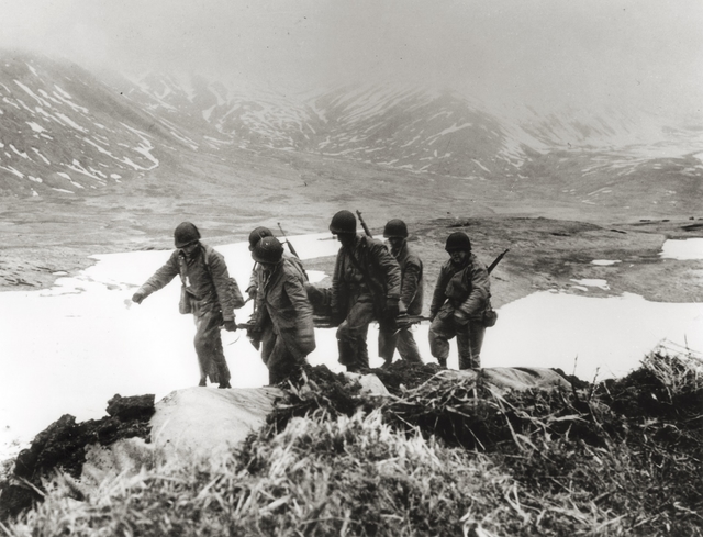 Six men with guns carry a person on a stretcher in a valley.