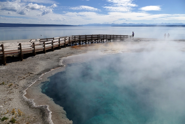 A visitor stands on a boardwalk near a hot spring and a lake.