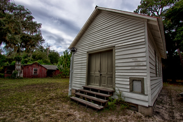 small white one room church, old red house in the background, under cloudy skies