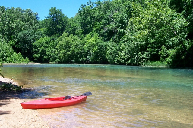 A red canoe on gravel bar with clear blue stream behind and green forest