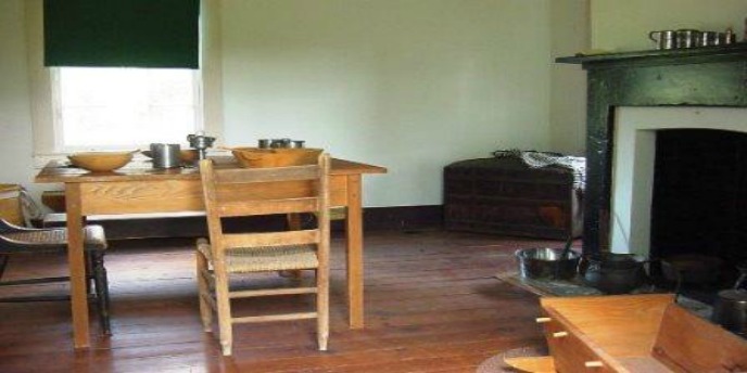 Room containing very simple wood table and chair, and baby cradle with fireplace in background