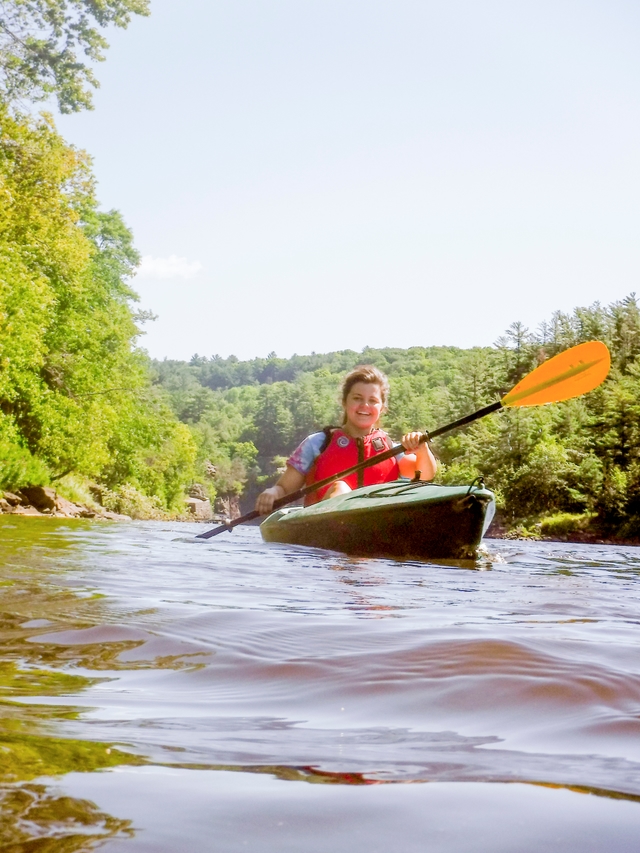 A woman paddles a kayak past cliffs on a forested river.