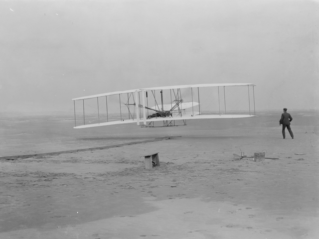 Orville takes to the air in the flyer for the first time as Wilbur assists.