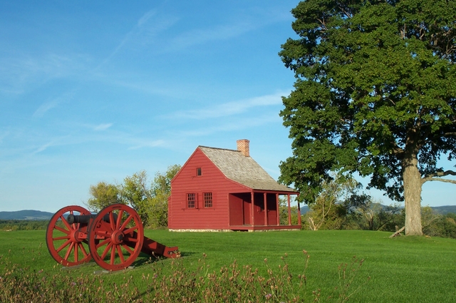 A small, red farmhouse sits beside a red cannon and a few green trees.