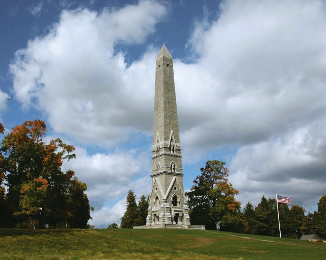 A narrow, stone obelisk on top of a green hill reaches into a partly cloudy sky.