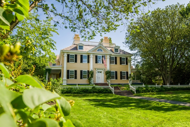 Yellow three-story mansion with symmetrical facade. Steps and large lawn in foreground. Framed by br