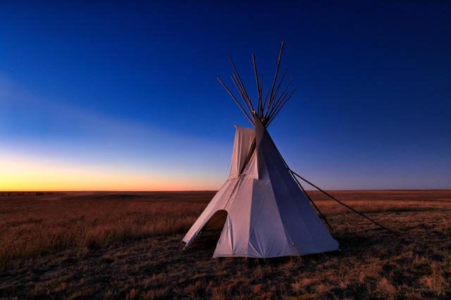 A lone tipi stands on a prairie landscape in predawn light.