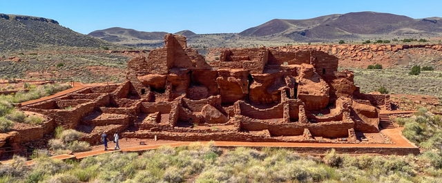 a large stone pueblo in a desert beneath mostly sunny skies with mountains in background