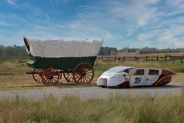 A Conestoga wagon and a solar race car stand on a road with a large adobe fort in the background