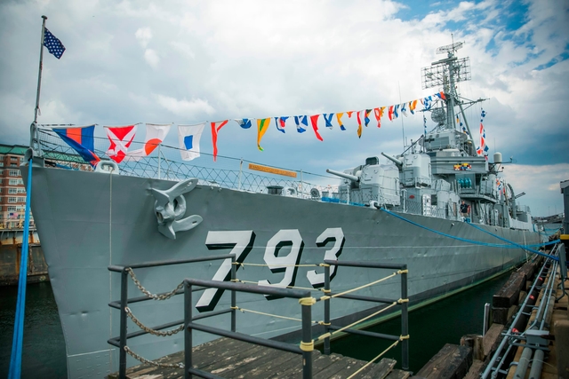 A gray naval destroyer sits next to a dock. It has "793" on the side and colorful signal flags.
