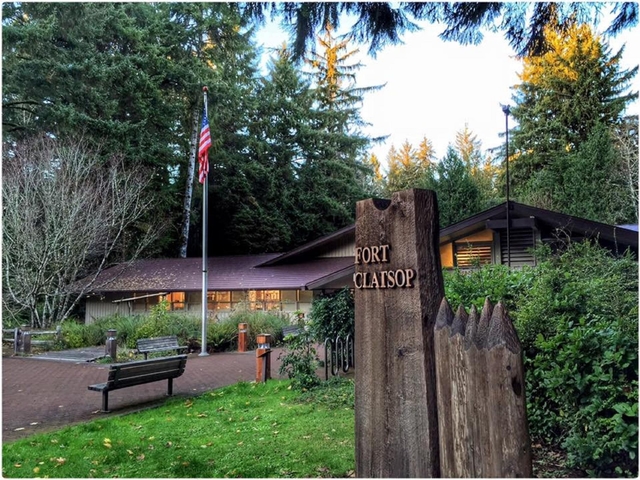 A wooden rectangular sign reading Fort Clatsop eclipsing a wide one story building and flag pole