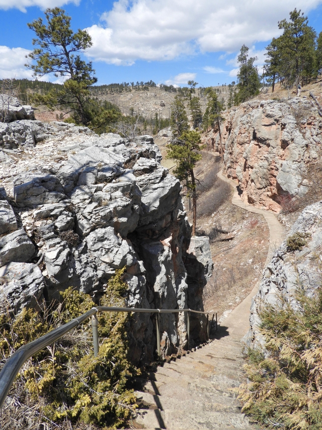 A paved path with steps heads down a hillside, with rock overhangs and pine trees nearby.