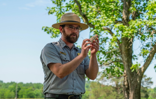 A park ranger holds up a specimen of limestone as he speaks outdoors.