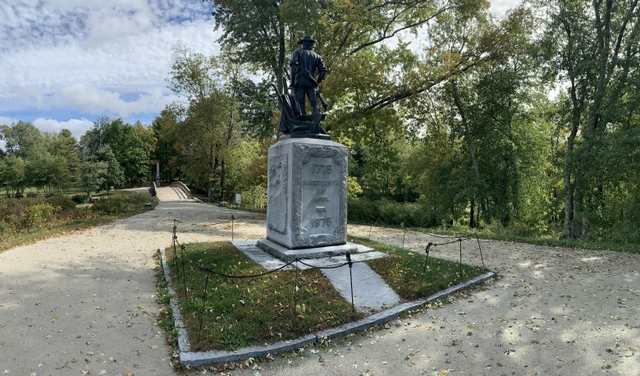 The state of the Minute man stands atop a stone base. The wooden North Bridge is seen in the distanc