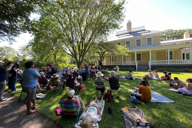 Crowd seated on a shaded lawn listening to an outdoor concert with yellow historic mansion
