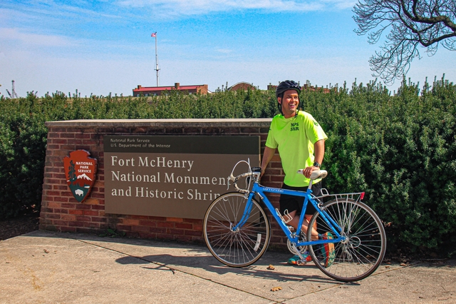Man in brought yellow shirt with a blue bicycle standing in front of a Fort McHenry identifier sign.