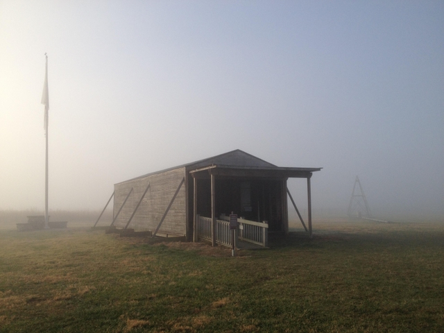 The fog lifts as the suns rays peek through the mist revealing a wood-frame hangar and flag pole.