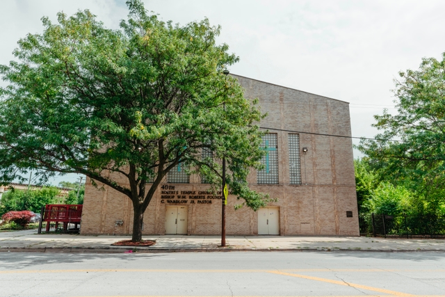 A multi-story tan brick church along a road. A tree stands in front.