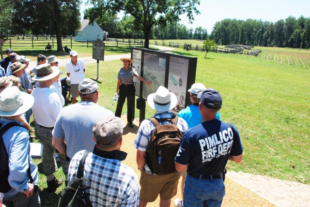 A uniformed park ranger gestures to an upright sign in front of a crowd of visitors.