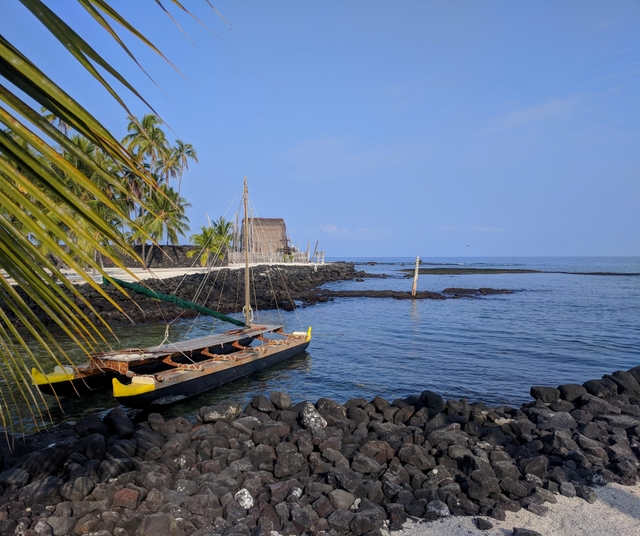 A double-hulled canoe sits in Keoneʻele Cove with Hale o Keawe in the background.