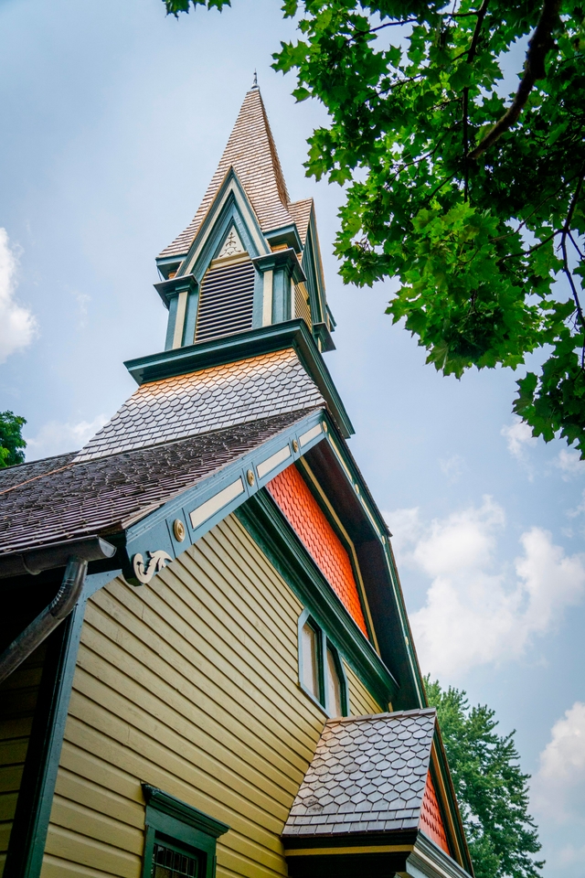 A tall, green and orange painted church steeple, against a blue sky, with a tree in the foreground.