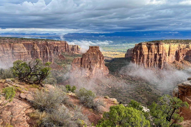 a wide sandstone monolith stands next to cliffs on a misty, cloudy day with green shrubs below