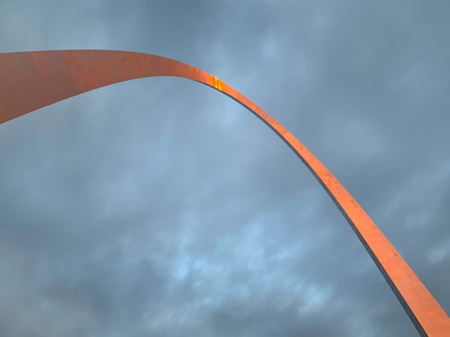 looking up at the Gateway Arch against a cloudy blue sky with a orange reflection at the top