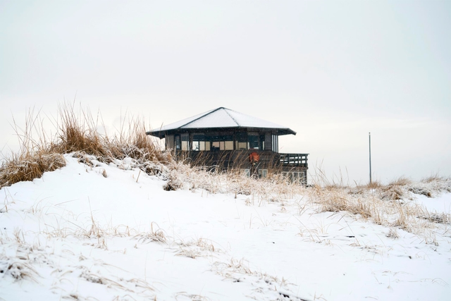 A brown hexagonal building covered in snow stands behind a beachside dune.