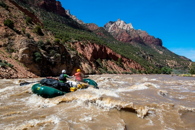 A green rubber raft floats over a rapid on a brown colored river in front of multi-colored mountains