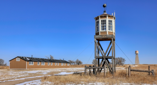 reconstructed barracks, guard and water towers surrounded by brown grass and shrubs.