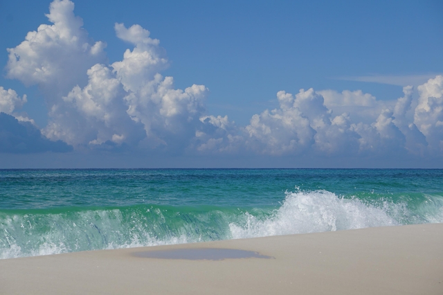Fluffy white clouds shadow overhead as blue-green waves crash against a white sand beach.