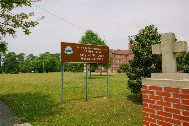 A brown sign reads "Campsite 4. City of St. Jude. March 24, 1965." Brick building in the background.