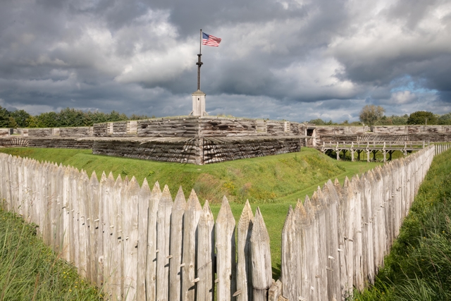 Puffy white clouds billow over log cabin-like walls. An American flag flies on a wooden pole